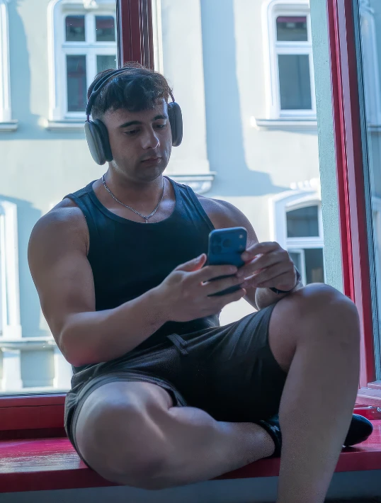Un ragazzo seduto sul davanzale di una finestra aperta, con le cuffie e il telefono in mano.